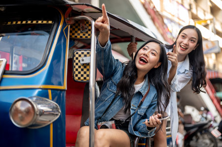 Two excited and pretty Asian girl tourists enjoy a ride in a tuk-tuk in Thailand, pointing at the view and having fun together while experiencing Thailand's transportation. Southeast Asia tourismの写真素材