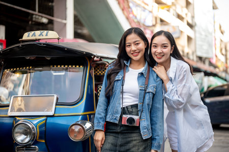 Two beautiful and cheerful Asian female tourists stand by a tuk-tuk on the street, enjoying their trip in Thailand and smiling at the camera. Thailand transportation, traveling, tourism, vacationの写真素材