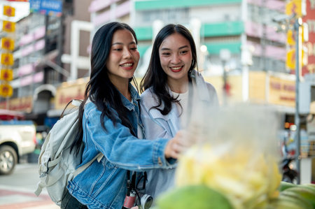 Two cheerful, cute Asian female tourists buy street food in Thailand, enjoying a summer stroll through Chinatown, traveling in Southeast Asia. food stall, street food, local marketの写真素材