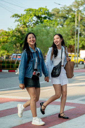 Two attractive, joyful young Asian women cross a crosswalk together while strolling through a city in Thailand on a sunny day, enjoy exploring the city.の写真素材
