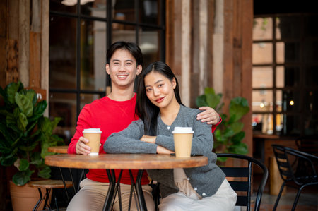A portrait of a sweet, happy young Asian couple in cozy clothes, sitting at an outdoor table of a coffee shop, enjoying a coffee date in autumn and sharing joyful moments together.の写真素材