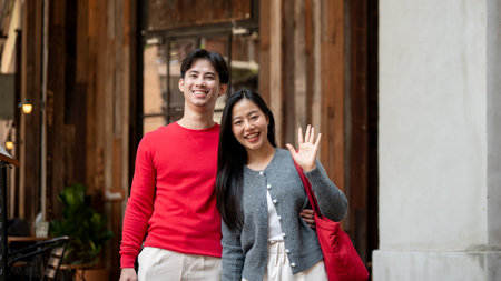 A portrait of a charming, lovely young Asian couple in cozy clothes, enjoying a weekend date as they stroll through the city and shop together, smiling and waving at the camera.の写真素材