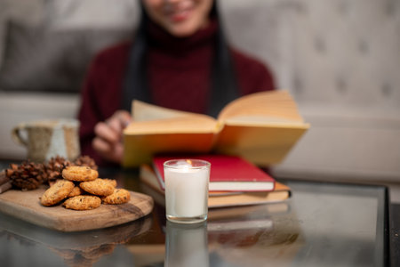 A wooden plate of cookies, a lit candle, stacked books, and a coffee mug on a coffee table. In the blurred background, a woman sits reading, creating a cozy Christmas at home atmosphere.の写真素材