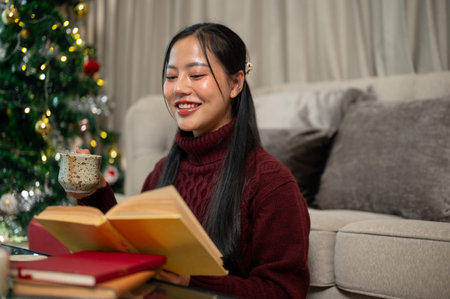 An attractive, happy Asian woman in a cozy red sweater sits in her living room, enjoying a book and some snacks during the Christmas holiday, spending the festive season at home alone.の写真素材