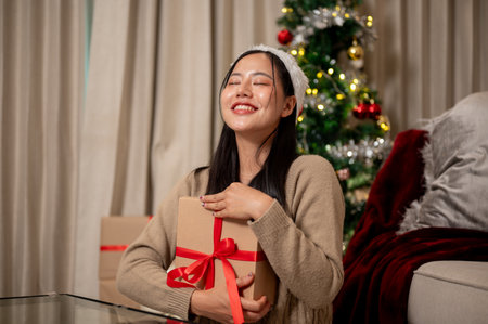 A cheerful, happy Asian woman sits in the living room, hugging her Christmas present box tightly, smiling with pure joy and happiness. Christmas holiday conceptの写真素材