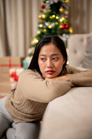 A sad, lonely Asian woman sits on the sofa, feeling bored, celebrating Christmas night alone at home. Christmas holiday conceptの写真素材