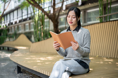 A charming Asian woman enjoys listening to music through her earphones and reading a book while sitting on a park bench, relaxed in the serene outdoor setting.の写真素材