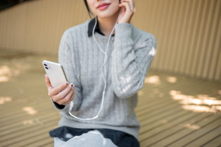 A cropped image of a woman in cozy clothes sitting on a park bench on a bright day, enjoys listening to music through her earphones, relaxing outdoors.の写真素材
