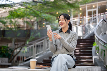 A beautiful, happy Asian woman in warm clothes sits outdoors on a bright day, using her smartphone with a smile. reading text messages, scrolling mobile apps, people and wireless technology conceptsの写真素材