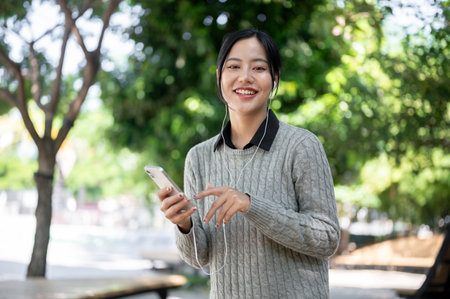 A charming young Asian woman in warm clothes enjoys listening to music through her earphones while walking along the footpath, taking in the green, vibrant surroundings.の写真素材