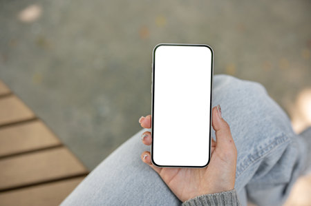 A close-up of a smartphone with a white screen mockup in a woman's hand, set against a blurred outdoor background. The woman is sitting on a park bench, using her smartphoneの写真素材