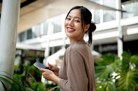 A portrait of a beautiful, positive Asian woman in casual clothes stands outdoors with a smartphone...