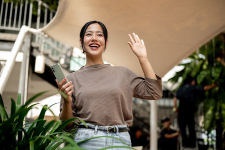 A beautiful, cheerful Asian woman in casual clothes smiles brightly and waves as she unexpectedly...