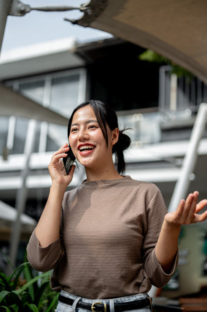 A cheerful Asian woman talks on her phone while walking outdoors. people, lifestyles, wireless technologyの写真素材