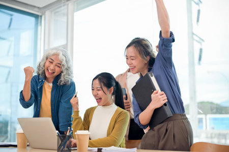 A group of cheerful, diverse Asian businesspeople celebrate their project success in the office, raising their fists in excitement while looking at a laptop screen. team building, teamwork, unityの写真素材