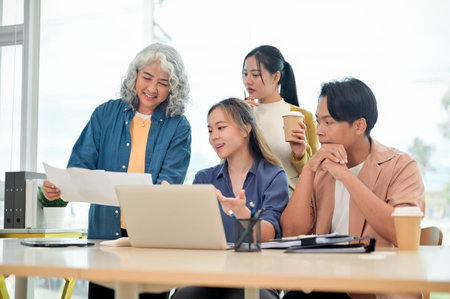 A generous senior Asian female boss assists her employees with a project, offering advice and helpful ideas while brainstorming together in a modern office. A startup team working together in office.の写真素材