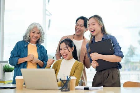 A cheerful Asian startup team celebrates their project success together in the office, looking at a laptop screen, showing their fists in excitement, and smiling with joy.の写真素材