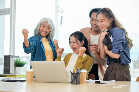 A cheerful Asian startup team celebrates their project success together in the office, looking at a laptop screen, showing their fists in excitement, and smiling with joy.の写真素材
