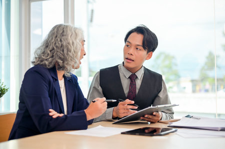 A young, smart Asian male businessman is discussing work with his senior female boss in a modern office. business meeting, consultation, lawyer, insuranceの写真素材