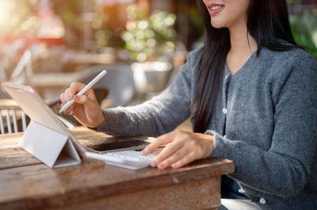 A cropped image of a beautiful Asian woman in a cozy sweater working remotely at a coffee shop in the city, using her digital tablet, with a smartphone and calculator resting on the table.の写真素材