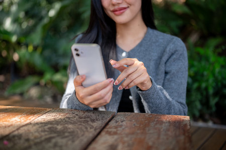 A close-up of an attractive Asian woman in a cozy grey sweater sitting at an outdoor cafe table in the city on a bright day, using her smartphone to scroll through social media.の写真素材