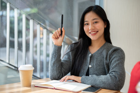 A portrait of a beautiful Asian woman in a cozy grey sweater sitting at a table in a coffee shop, holding a pen and smiling at the camera, with a book, smartphone, and coffee cup placed on the table.の写真素材