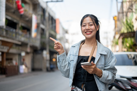 A beautiful, smiling Asian female tourist in a denim jacket holding her smartphone, standing by the street and pointing her finger upward as she recommends something.の写真素材