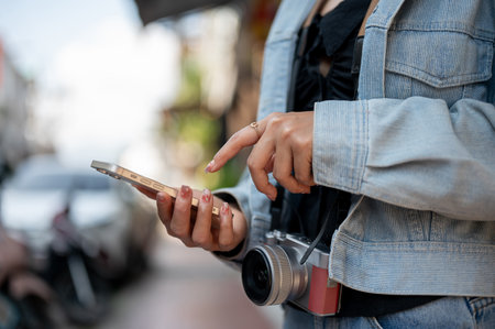 A close-up of a female tourist in a denim jacket using her smartphone to check directions on an app while exploring the city, walking down the street.の写真素材
