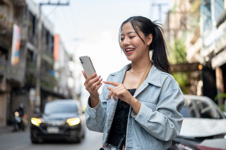 An attractive, happy Asian female tourist in a denim jacket using her smartphone to check directions on an app while exploring the city, walking down the street.の写真素材