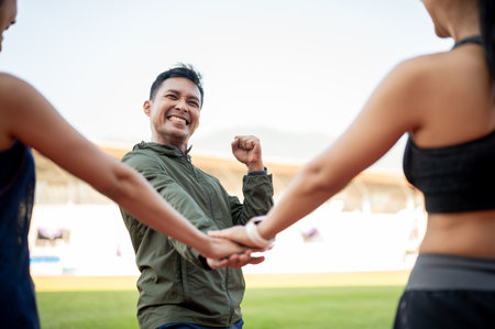 A group of cheerful Asian runners puts their hands together in a gesture of encouragement before running at a stadium in the morning. friendship, sport, lifestylesの写真素材