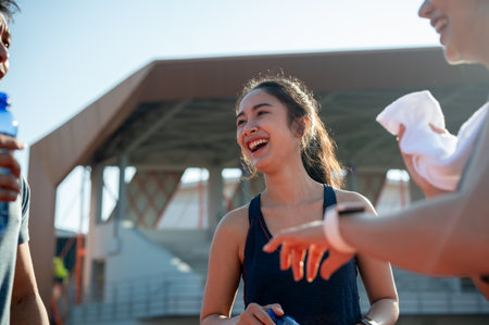 A happy Asian female athlete in sportswear enjoys a conversation with her friends while resting after training at a stadium on a sunny day. people and sport conceptsの写真素材