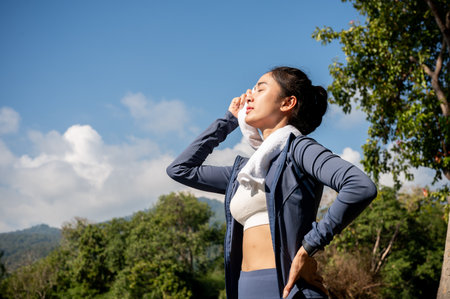A tired, sweaty Asian woman in sportswear wipes sweat from her face with a towel, feeling exhausted and hot while exercising in a park on a sunny summer day. people and sport conceptsの写真素材