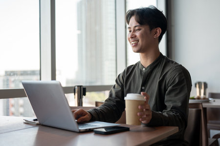A positive, handsome Asian businessman enjoying a hot drink while working on his laptop, working remotely from a coffee shop in a modern building with a view of skyscrapers.の写真素材