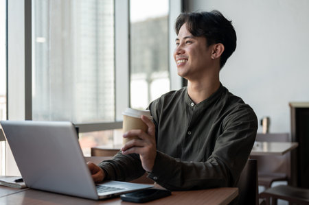 A positive, handsome Asian businessman enjoying a hot drink while working on his laptop, working remotely from a coffee shop in a modern building with a view of skyscrapers.の写真素材