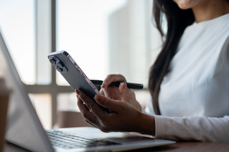 A close-up of a beautiful Asian businesswoman touching her smartphone screen, using her smartphone while working in a modern office in a skyscraper.の写真素材