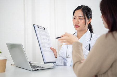A confident Asian female doctor showing a medical paper to a patient while discussing the treatment plan during a consultation in the examination room at the hospital.の写真素材