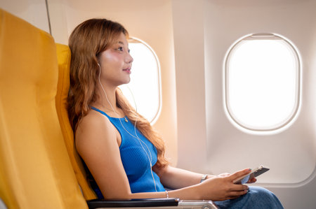 A side view of a relaxed, happy Asian female passenger sitting at a window seat, enjoying listening to music through her earphones during the flight. people, airplane, traveling, lifestyleの写真素材