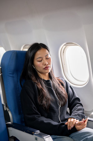 A relaxed and calm Asian female passenger falls asleep while listening to music through her earphones during the flight, sitting at a window seat, traveling by plane.の写真素材
