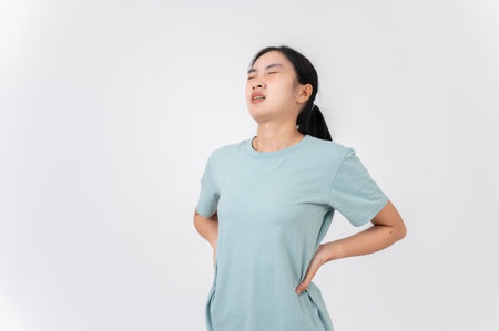 A tired, uncomfortable young Asian woman touching her back, suffering from back pain, period cramps or office syndrome, standing against on a white studio background. health care conceptの写真素材