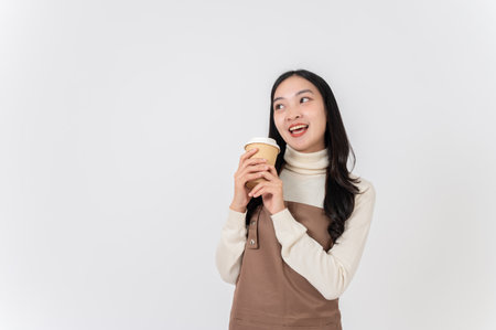 A cheerful, smiling Asian female barista or coffee shop waitress in a brown apron holding a hot drink cup, standing on a white studio background with a copy space. small business owner conceptの写真素材