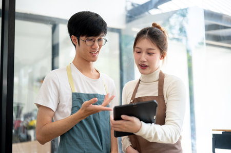 Two Asian coffee shop staff members are discussing the menu on a tablet while working in their coffee shop during the daytime. small business, part-time job, business ownerの写真素材