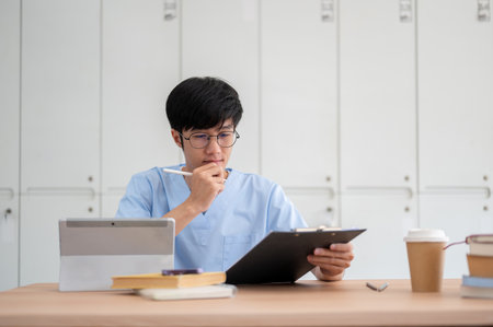 A smart Asian male medical student in scrubs, focused on reviewing medical cases on a clipboard at his desk in a library or classroom. medical and people conceptsの写真素材