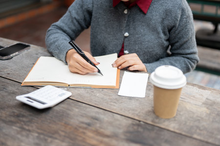 A cropped image of a woman writing or taking notes in a book while sitting at a table outdoors, planning her monthly budgets, calculating her expenses and bills.の写真素材
