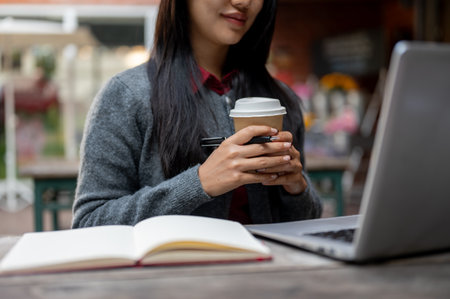 A close-up of a positive Asian female student studying online on her laptop while sitting at an outdoor cafe table, sipping a hot drink and concentratively on the lesson on the laptop.の写真素材