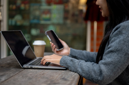 A close-up of a woman holding her smartphone while working on her laptop, typing on the laptop...