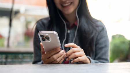 A cropped image of a positive Asian woman enjoys listening to music on her earphones and using her...