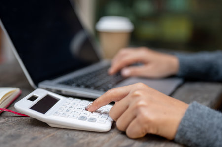 A close-up of a woman using a calculator while working on her laptop at a table outdoors, calculating expense, working on her business financial.の写真素材