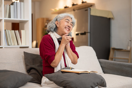 A happy Asian retired senior woman daydreams about her happiness while writing in her diary on a sofa in the living room. domestic life, leisure, wellbeing lifestyleの写真素材
