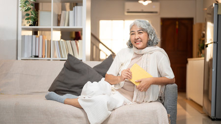 A happy Asian retired senior woman relaxes on a sofa, contemplating about happy life while reading a book. leisure activity, literature, domestic lifeの写真素材