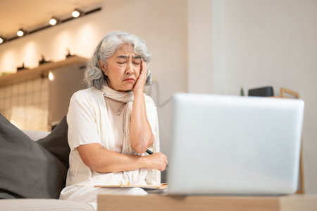A tired Asian retired woman has a headache while working on her laptop, sitting on a sofa in the living room. people, technology, lifestyleの写真素材